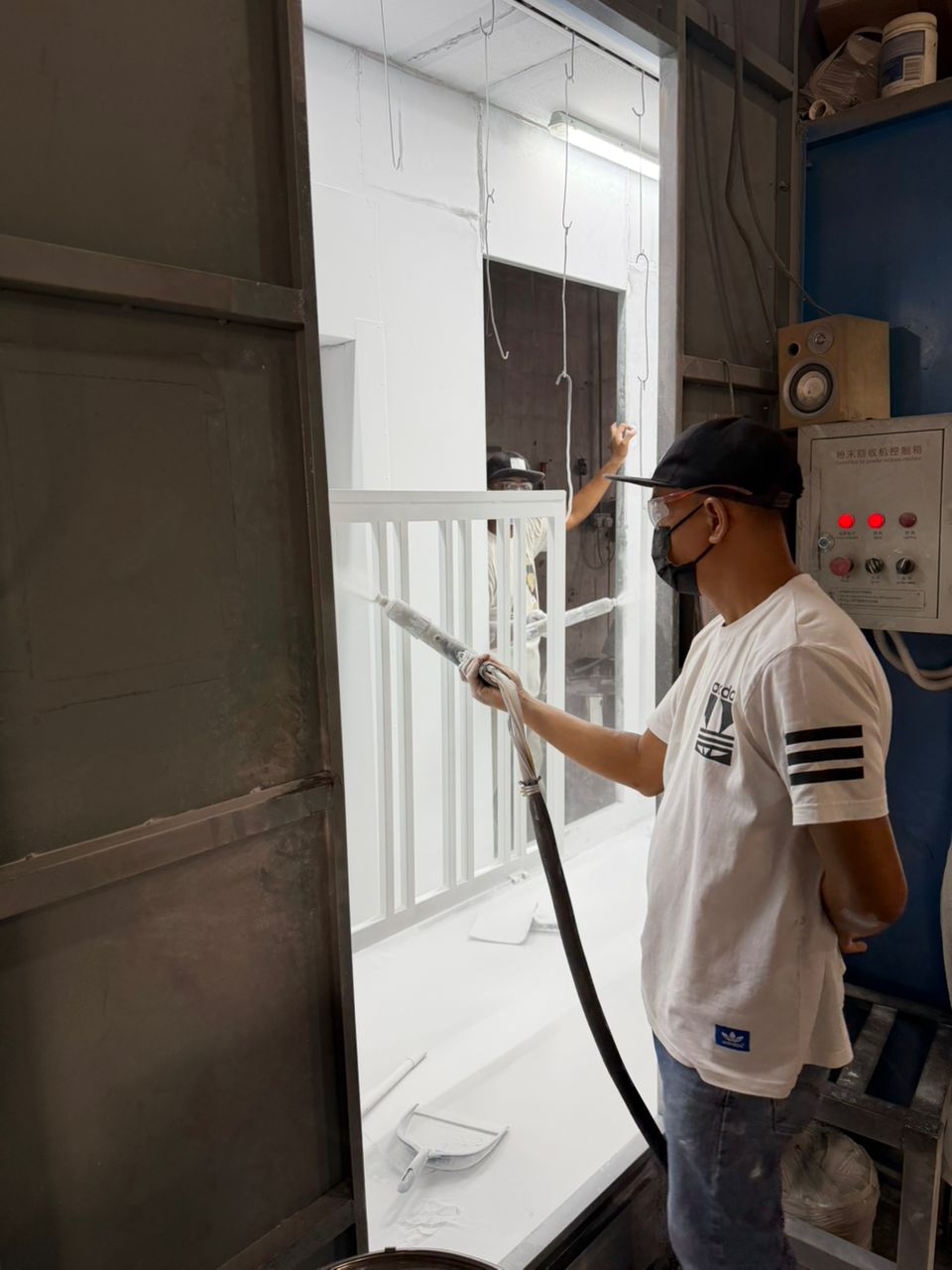 Worker in a mask spraying white powder coat onto a grille inside the spray booth